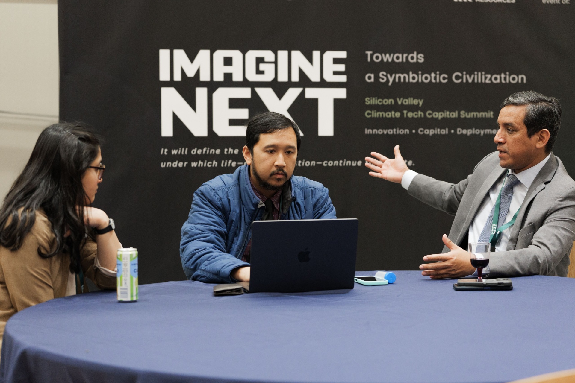 Three delegates in animated conversation around a round table beneath the Imagine Next 'Towards a Symbiotic Civilization' Silicon Valley Climate Tech Capital Summit backdrop during Capital Day at UC Law SF
